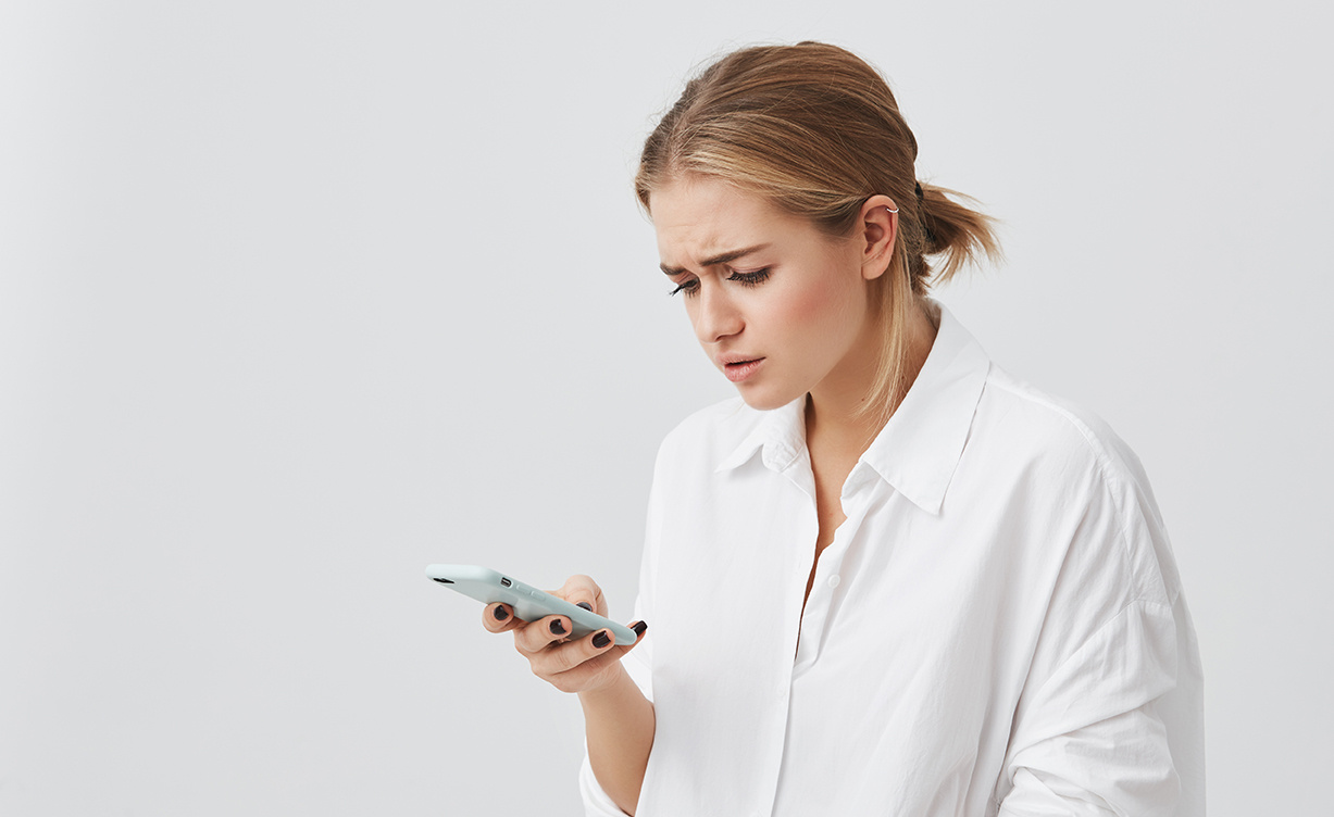 people and modern communication. studio portrait of worried pretty blonde girl reading urgent text message via smart phone. young female looking confused holding cell phone in her hand.