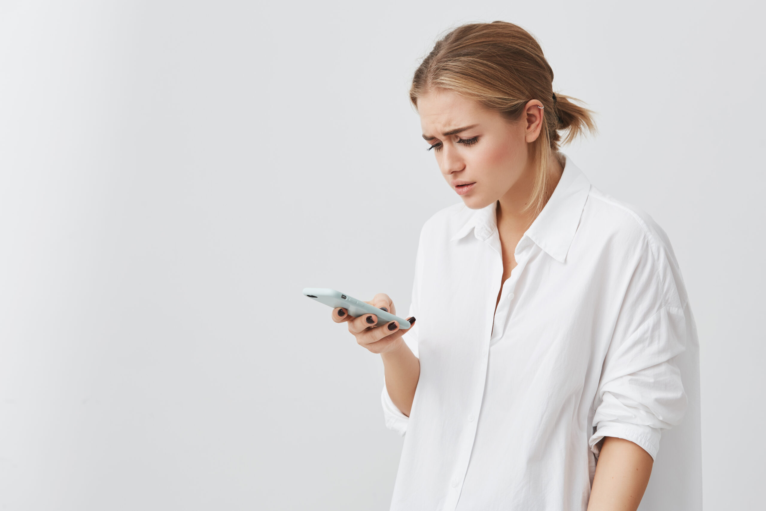 people and modern communication. studio portrait of worried pretty blonde girl reading urgent text message via smart phone. young female looking confused holding cell phone in her hand.